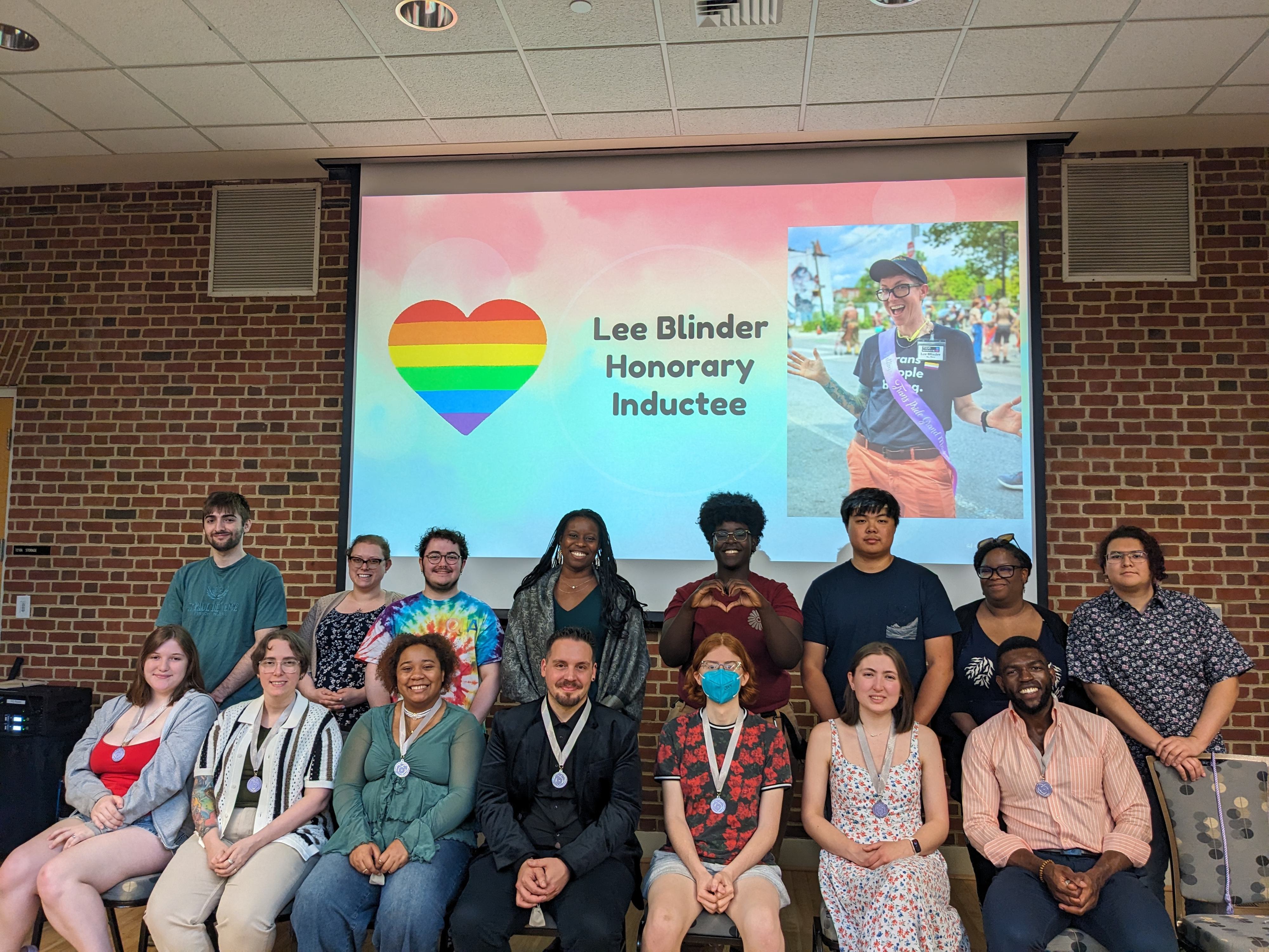 Group photo of Lavender Leadership Honor Society inductees, some wearing medals, in front of a slide projector that reads "Lee Blinder, honorary inductee" with a rainbow heart
